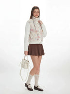 Side view of a woman modeling a floral print fleece bomber jacket with cable knit sleeves and a brown pleated skirt.