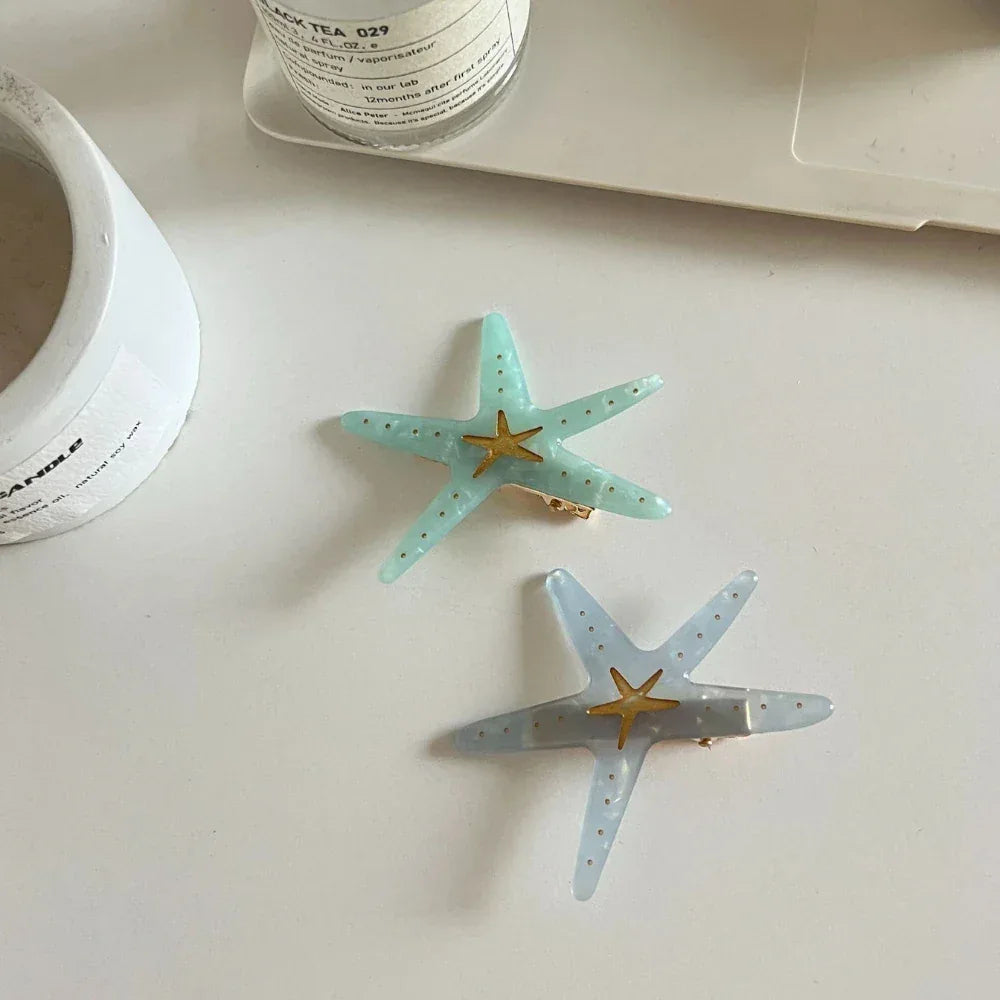 Two starfish hair clips, one light blue and one light grey, are shown on a white surface with a candle and laptop in the background.