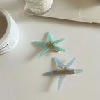 Two starfish hair clips, one light blue and one light grey, are shown on a white surface with a candle and laptop in the background.