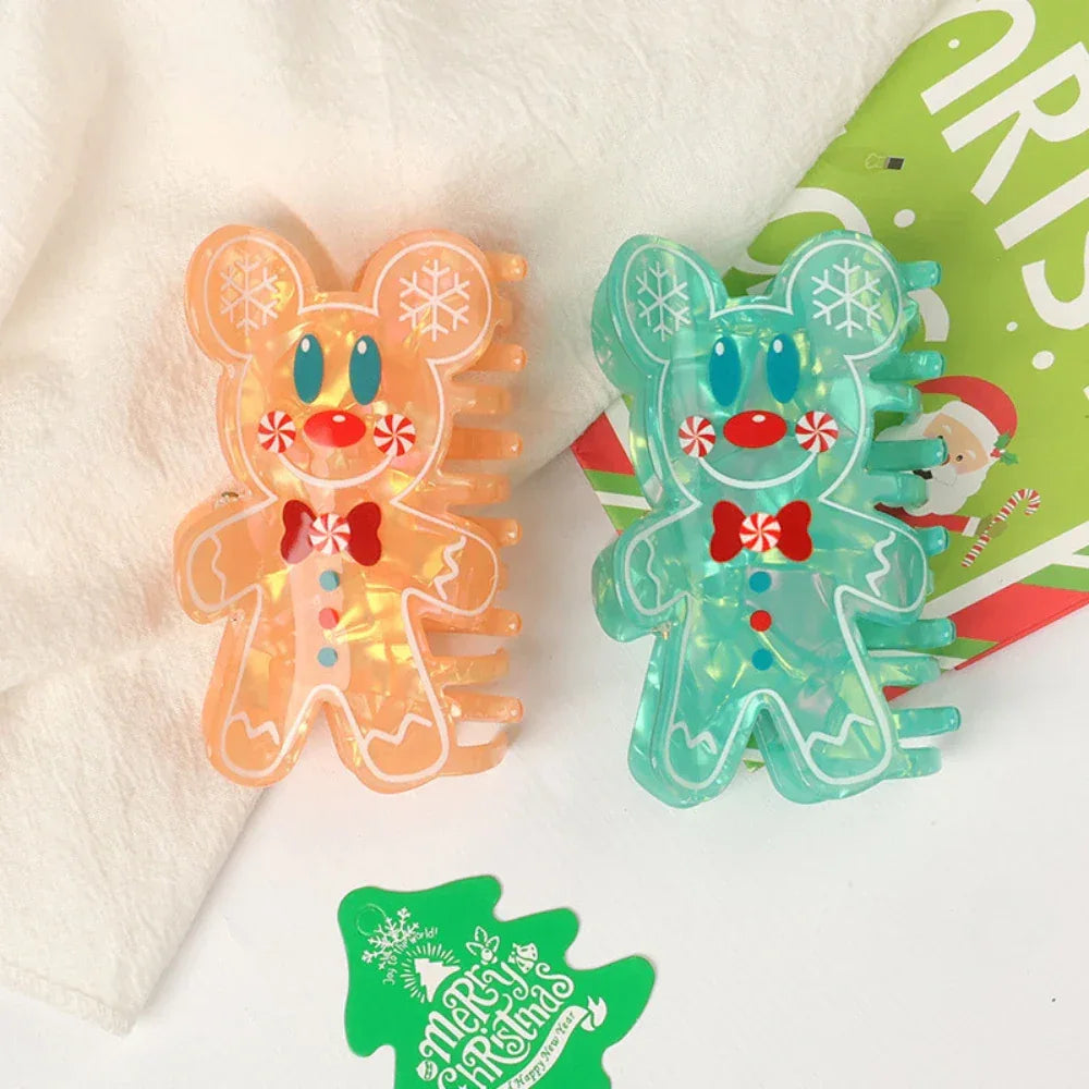 Two gingerbread man hair claw clips, one orange and one teal, displayed side-by-side on a white cloth with a Christmas-themed card in the background.