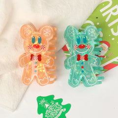 Two gingerbread man hair claw clips, one orange and one teal, displayed side-by-side on a white cloth with a Christmas-themed card in the background.