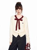 Close-up of a cream-colored cardigan with a burgundy bow and trim, featuring a small cat embroidery on the chest.