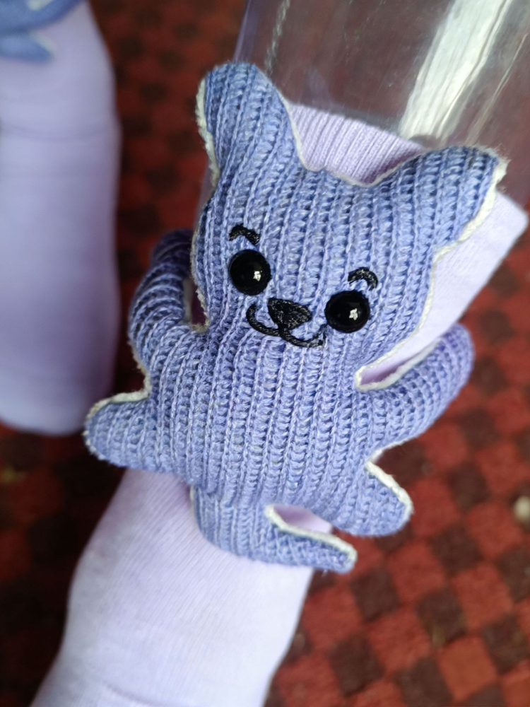 Detailed view of a blue knitted bear embellishment with button eyes and embroidered facial features, attached to a light purple sock.