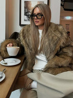 Woman in a brown faux fur jacket and beige pants, seated at a table with a cup of coffee.
