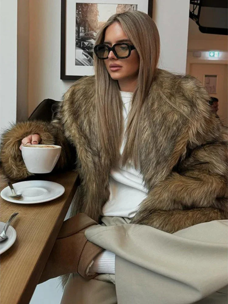 Woman in a brown faux fur jacket and beige pants, seated at a table with a cup of coffee.
