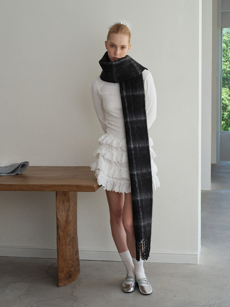 Model wearing a long, black and grey plaid scarf draped over a white long-sleeved shirt and a white ruffled skirt. Main image