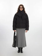 Model wearing a black puffer jacket with an attached scarf and a grey maxi skirt, holding a black tote bag.
