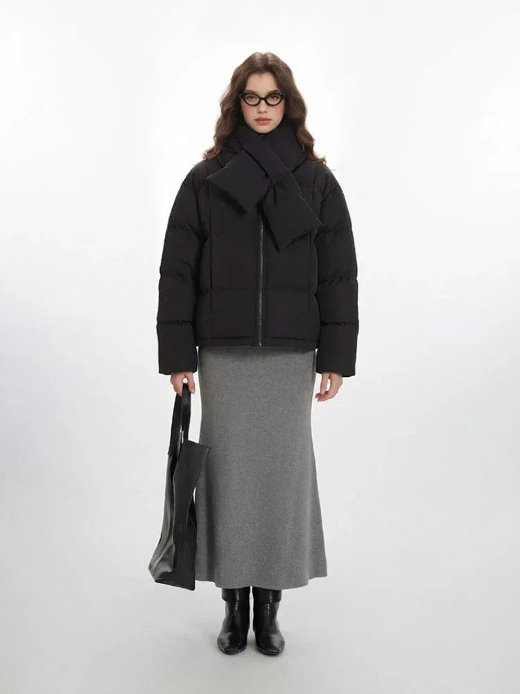 Model wearing a black puffer jacket with an attached scarf and a grey maxi skirt, holding a black tote bag.