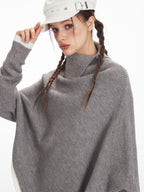 Close-up of a woman wearing a grey asymmetrical knit poncho with a turtleneck, adjusting her white cap