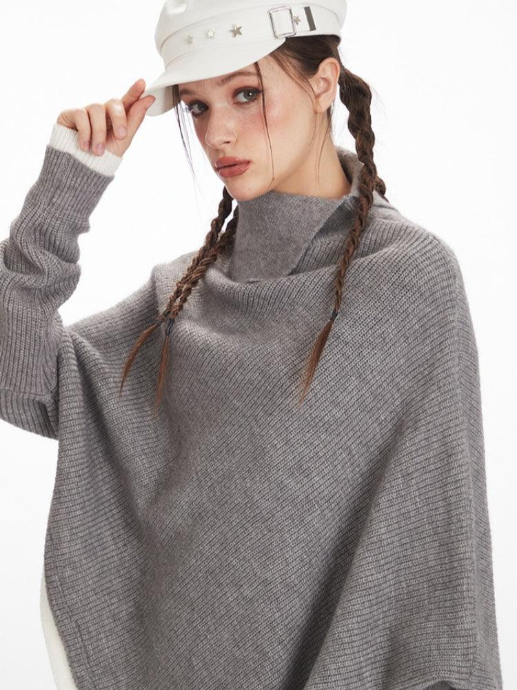 Close-up of a woman wearing a grey asymmetrical knit poncho with a turtleneck, adjusting her white cap