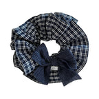A close-up, overhead shot of a large scrunchie with a navy and white plaid pattern and a dark blue textured bow. A small white tag with black text is visible on the bow.