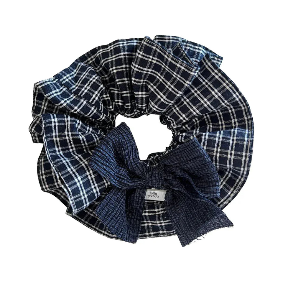 A close-up, overhead shot of a large scrunchie with a navy and white plaid pattern and a dark blue textured bow. A small white tag with black text is visible on the bow. Main image