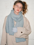 Model wearing a beige oversized coat with a light blue scarf and matching gloves. She is also wearing glasses.