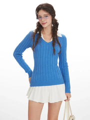 Woman wearing a blue cable knit V-neck sweater with a white collar and a white pleated skirt, posing with her hand on her hip, front view.
