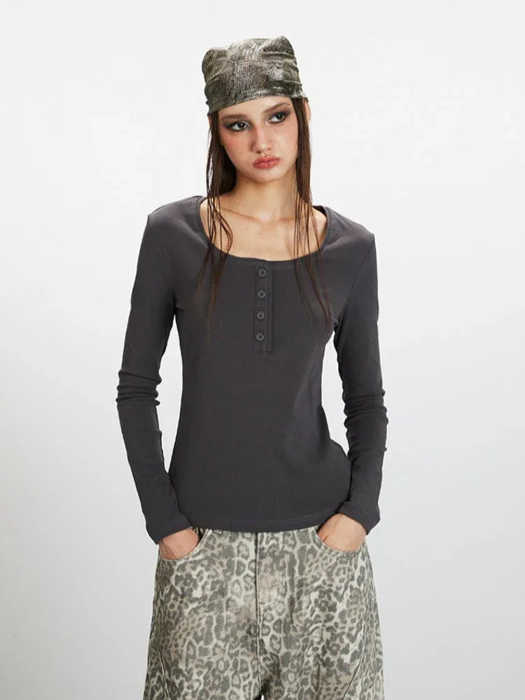Front view of a woman wearing a dark grey long-sleeve top with a square neckline and button details, paired with wide-leg grey leopard print pants. She is holding a dark handbag and wearing a patterned headscarf.