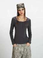 Front view of a woman wearing a dark grey long-sleeve top with a square neckline and button details, paired with wide-leg grey leopard print pants. She is holding a dark handbag and wearing a patterned headscarf.