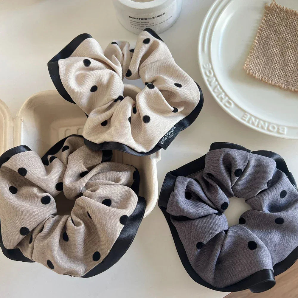 Three polka dot scrunchies, two beige and one gray, in a white container and on a white surface