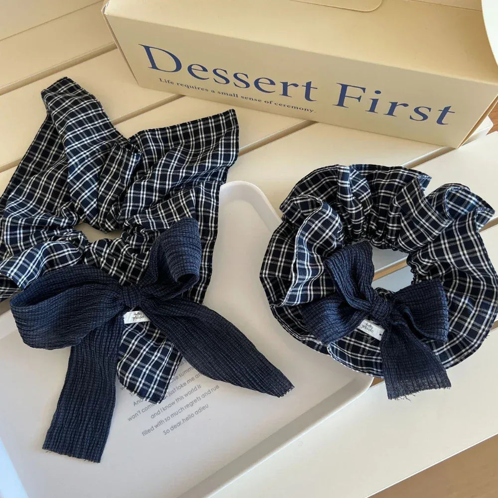 Two scrunchies with navy and white plaid patterns and dark blue textured bows are placed on a white tray next to a box with the text 'Dessert First'.