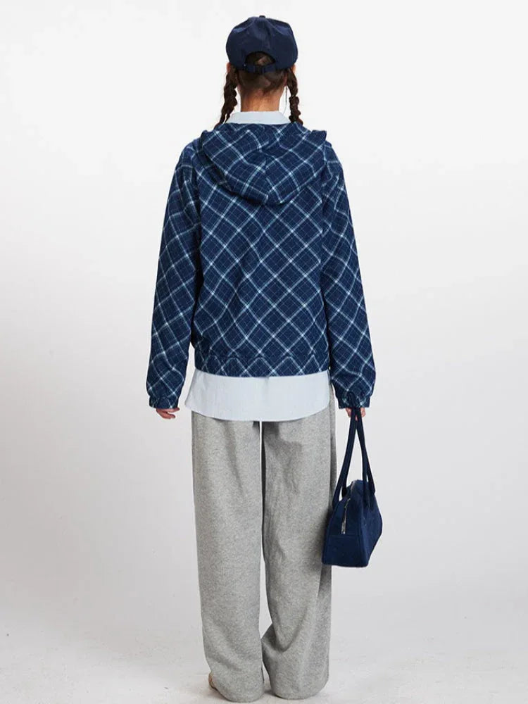 Back view of a model wearing a blue plaid hooded jacket, light blue collared shirt, grey sweatpants, and a navy blue cap, holding a navy blue handbag.