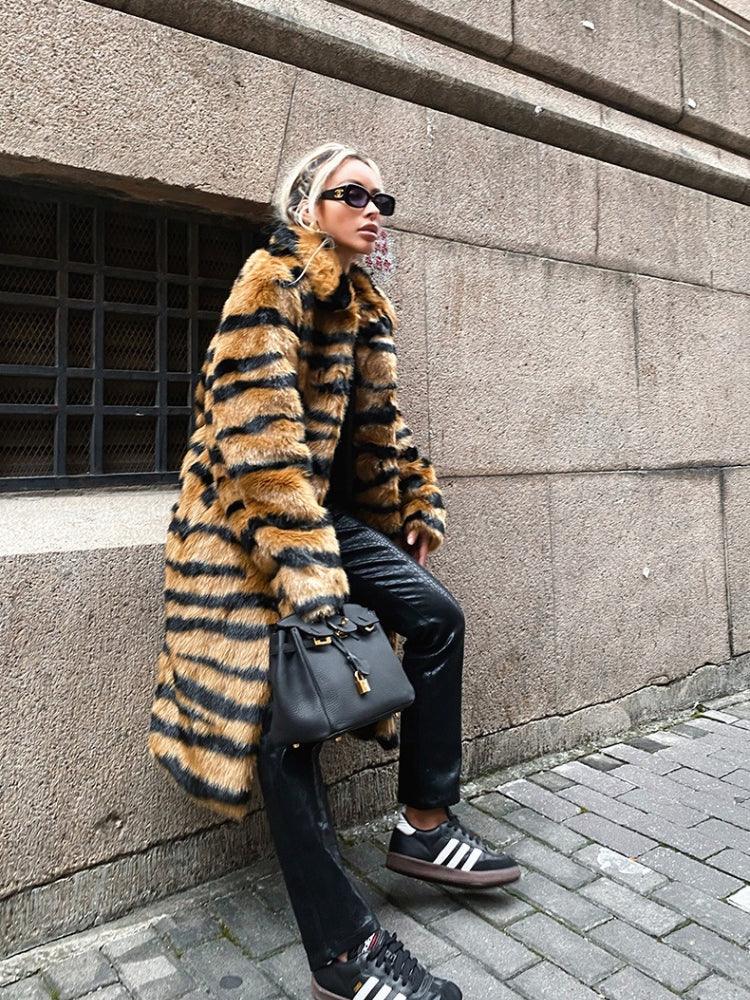 Full body shot of a woman in a faux fur tiger stripe coat, black pants, and sneakers, posing outdoors.