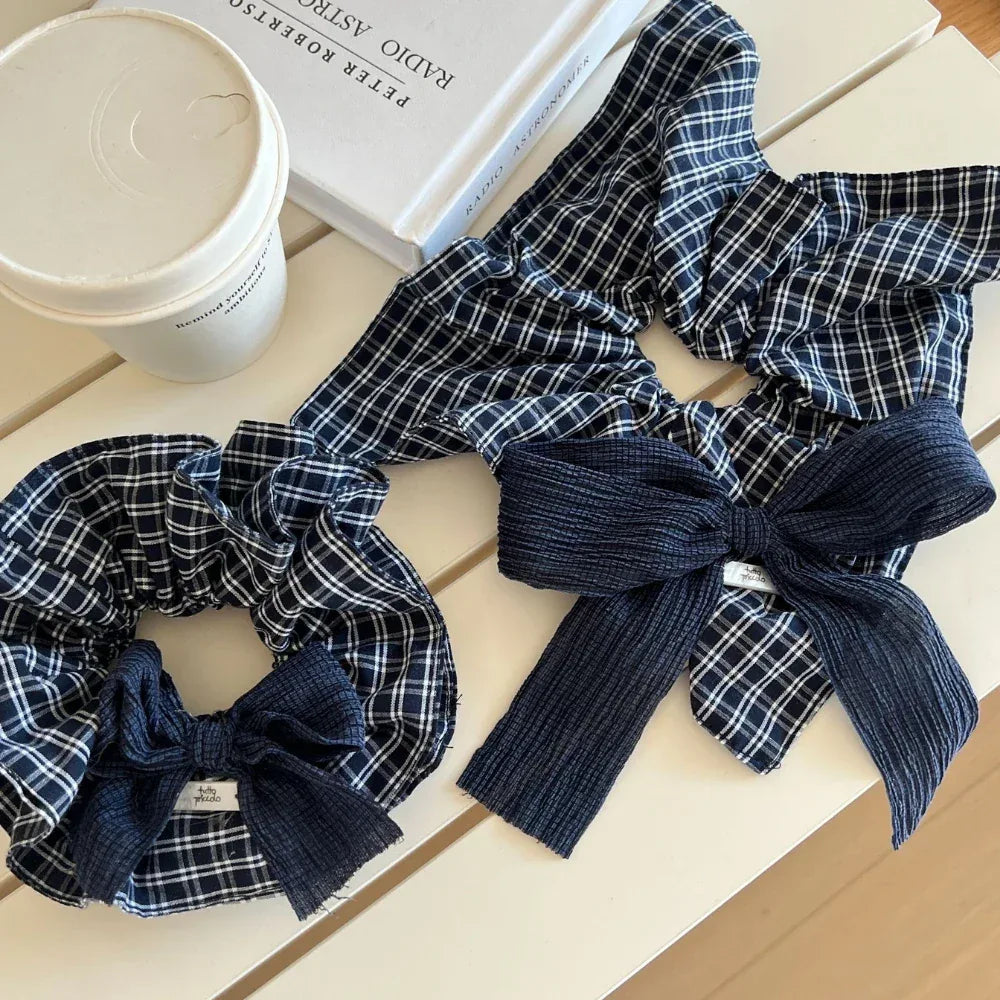 Two scrunchies with navy and white plaid patterns and dark blue textured bows are on a white table next to a white coffee cup and a book.