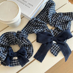 Two scrunchies with navy and white plaid patterns and dark blue textured bows are on a white table next to a white coffee cup and a book.