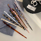 A variety of patterned hair sticks scattered on a grey fabric surface, next to a black dish with a pearl necklace.