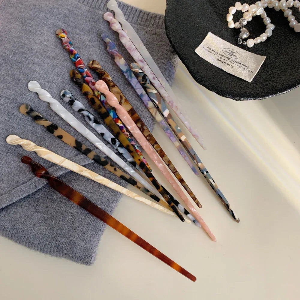 A variety of patterned hair sticks scattered on a grey fabric surface, next to a black dish with a pearl necklace.