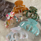A close-up view of four acetate claw hair clips in different marbled patterns: amber, green, multi-colored, and light blue. They are laid out on a textured white surface with a book in the background.