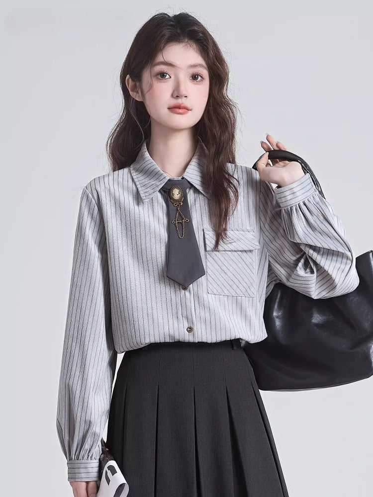 Woman wearing a grey pinstripe collared shirt with a dark tie and brooch, a black pleated skirt, and holding a black handbag.
