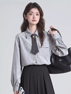 Woman wearing a grey pinstripe collared shirt with a dark tie and brooch, a black pleated skirt, and holding a black handbag.