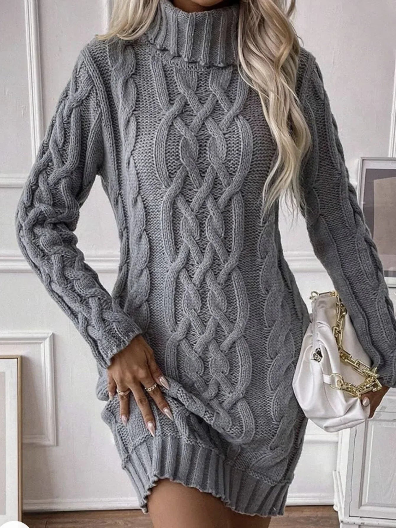 Woman wearing a grey cable knit turtleneck mini dress, adjusting the hem, and holding a white handbag.