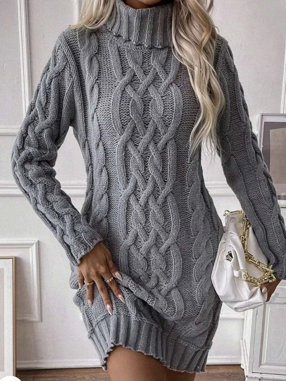 Woman wearing a grey cable knit turtleneck mini dress, adjusting the hem, and holding a white handbag.