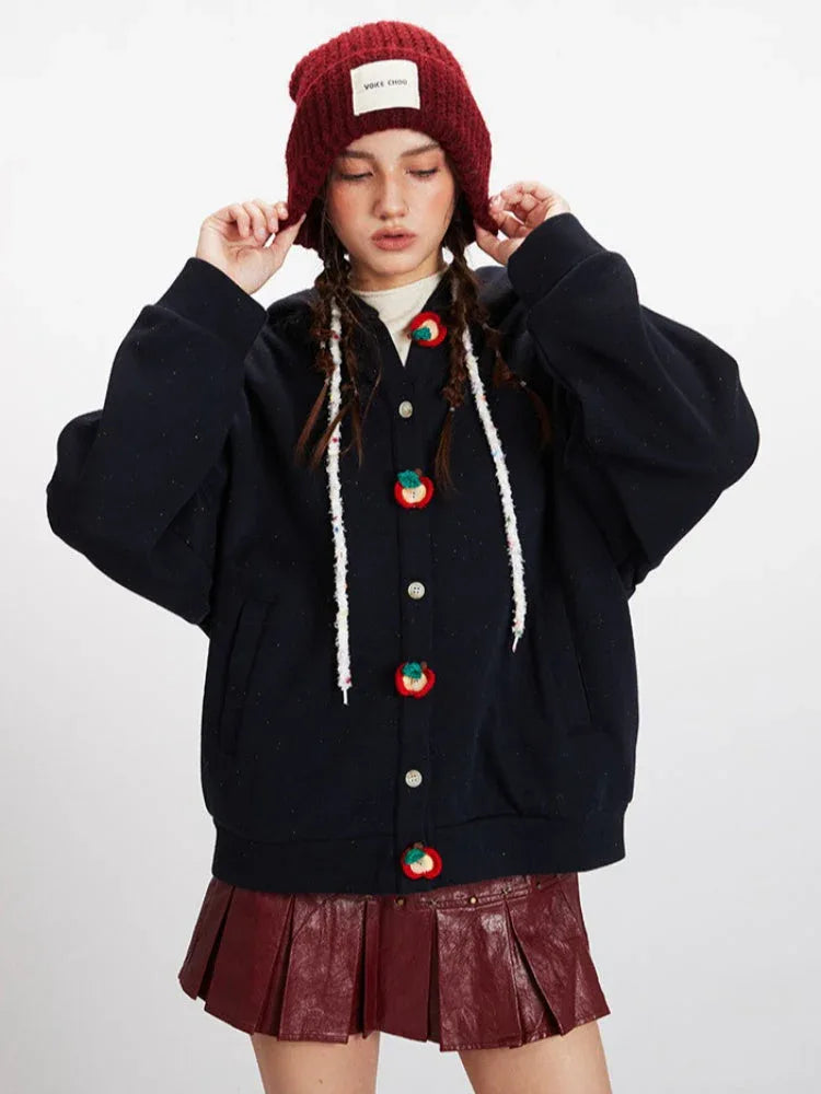 Front view of a woman wearing a dark blue hooded cardigan with colorful fruit buttons, adjusting her red beanie.