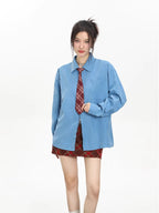 Model wearing an oversized blue collared shirt with a red plaid tie and a red plaid mini skirt, adjusting the zipper.