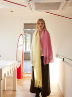 Woman wearing a two-tone yellow and pink fringed scarf draped over a white crochet top and brown wide-leg pants, standing in a hallway with a red mirror.