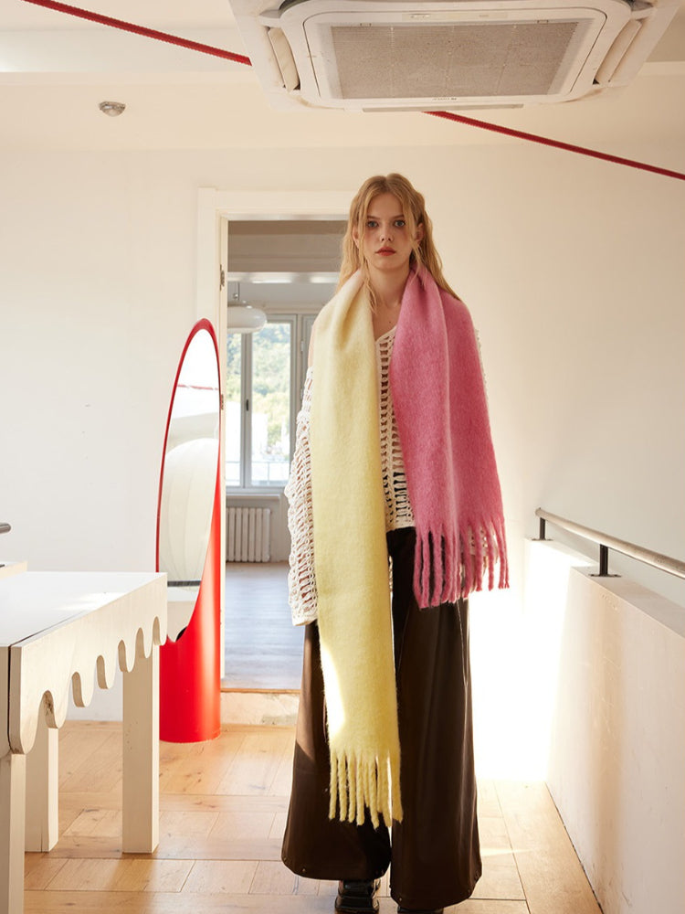 Woman wearing a two-tone yellow and pink fringed scarf draped over a white crochet top and brown wide-leg pants, standing in a hallway with a red mirror.