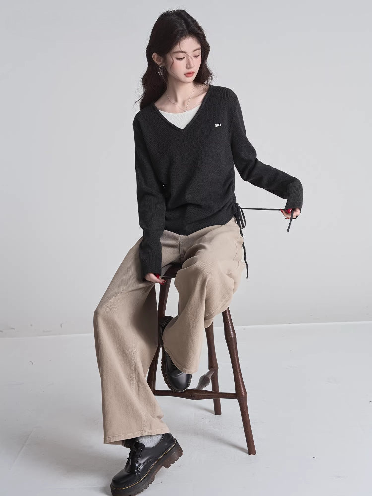 Full body shot of a woman sitting on a stool, wearing a dark grey v-neck sweater, white undershirt, and wide-leg tan pants.