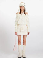 Model wearing a cream off-the-shoulder knit sweater and a matching mini skirt, with an ear flap hat and pink handbag.