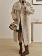 Woman wearing a long beige fuzzy fleece coat with a notched lapel and double-breasted closure, holding a tan and black handbag, paired with brown boots.