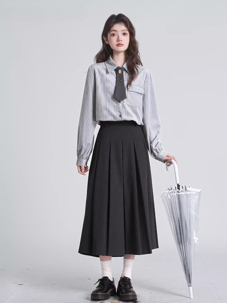 Full-body shot of a woman wearing a grey pinstripe collared shirt, black pleated skirt, and holding a clear umbrella.