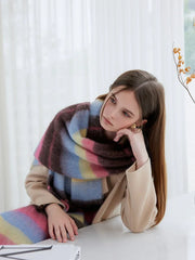 Woman leaning on a table, wearing a colorful striped scarf and a beige coat, looking to the side.