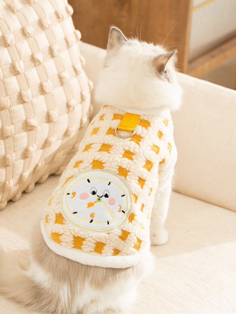 A fluffy white cat with blue eyes sits on a couch, facing away from the camera. The cat is wearing a yellow and white patterned fleece vest with a clock graphic on the back.
