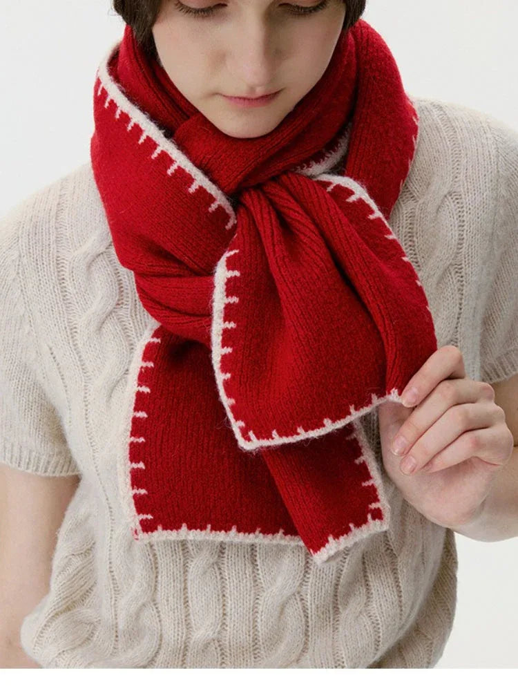 Woman adjusting a red knit scarf with white contrast stitching over a cream cable knit sweater.