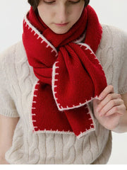 Woman adjusting a red knit scarf with white contrast stitching over a cream cable knit sweater.