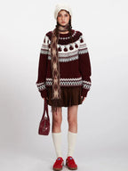 Woman wearing a maroon and white knit sweater with an apple Fair Isle pattern, brown corduroy skirt, white beanie, patterned scarf, white socks, and red sneakers.