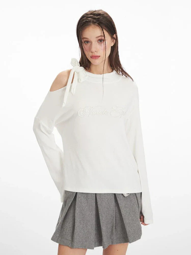 Front view of a woman wearing a white long-sleeved top with one shoulder exposed and a grey pleated skirt.