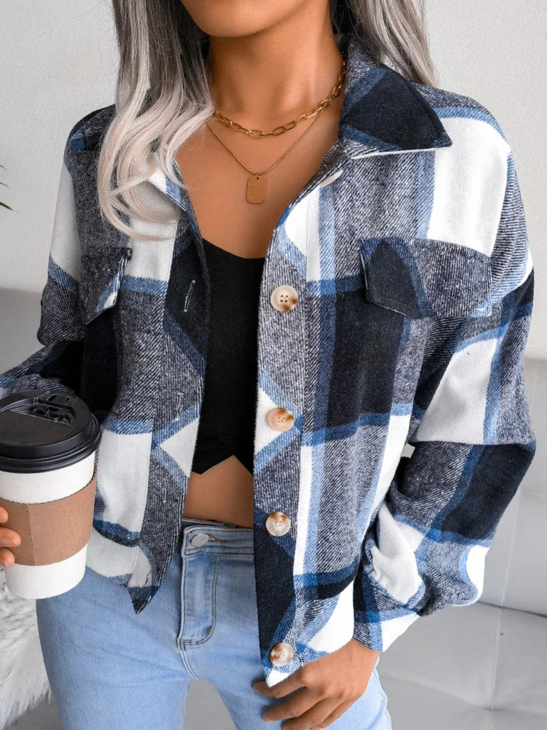Woman wearing a blue, white, and black plaid flannel shirt jacket over a black crop top and light wash jeans, holding a disposable coffee cup.