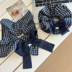 Two scrunchies with navy and white plaid patterns and dark blue textured bows are on a white table next to a laptop and a small print.