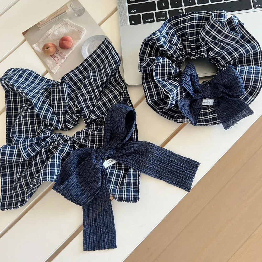 Two scrunchies with navy and white plaid patterns and dark blue textured bows are on a white table next to a laptop and a small print.